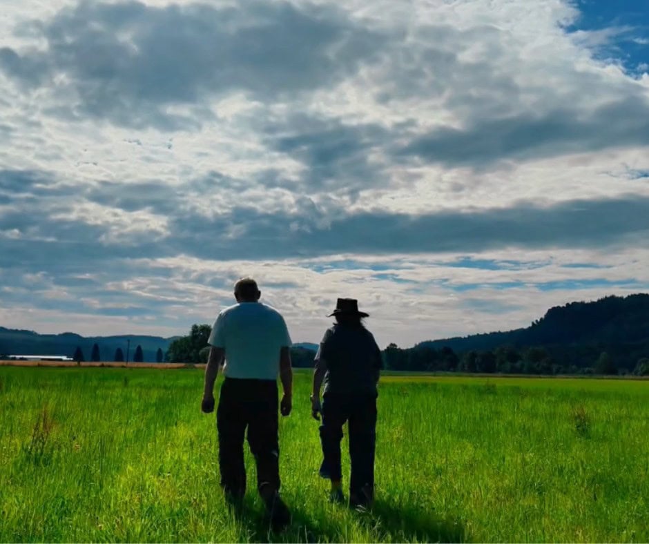 Two people walking through a lush green field under a cloudy sky.